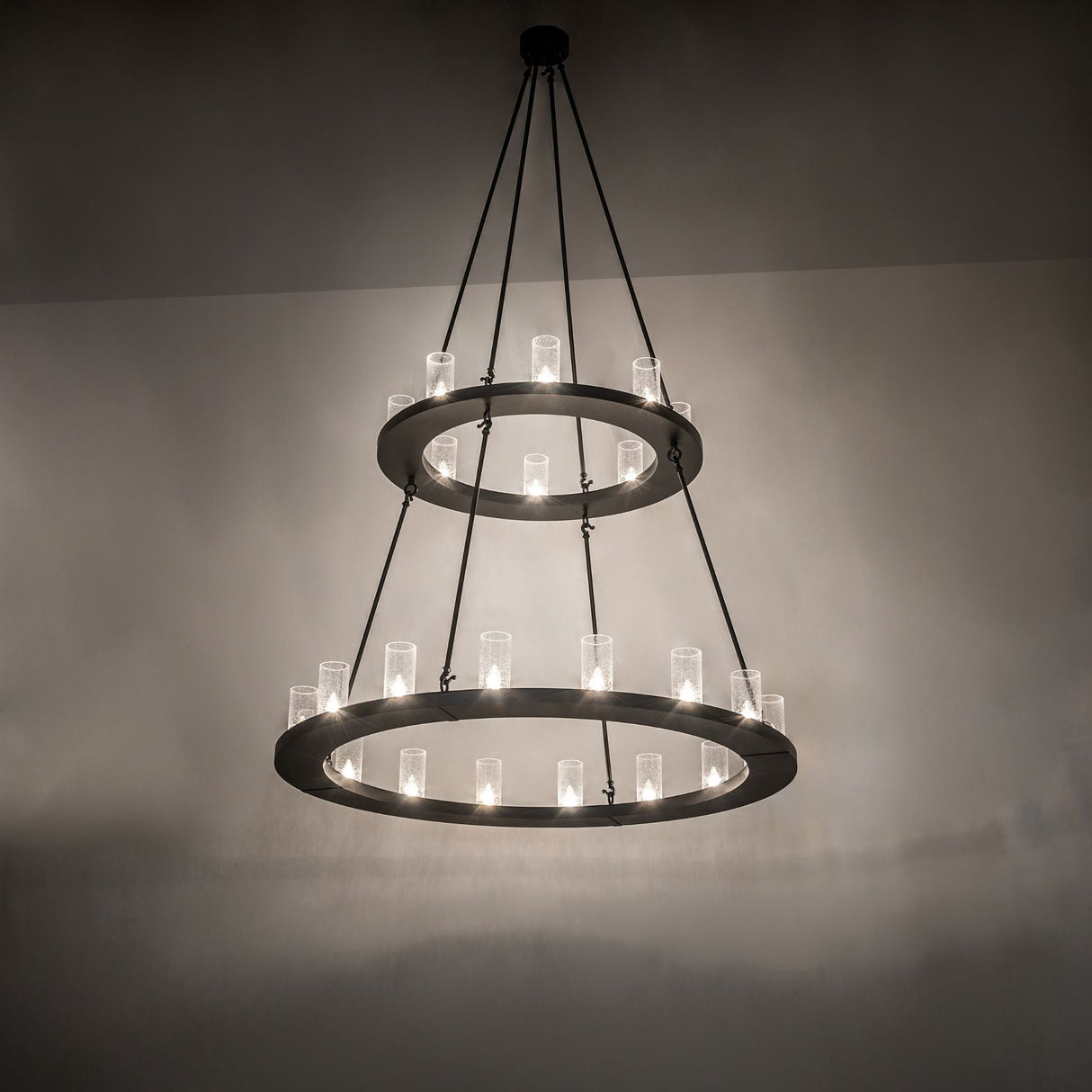 Ambient view of lit two-tier black chandelier with seeded glass cylinder shades.