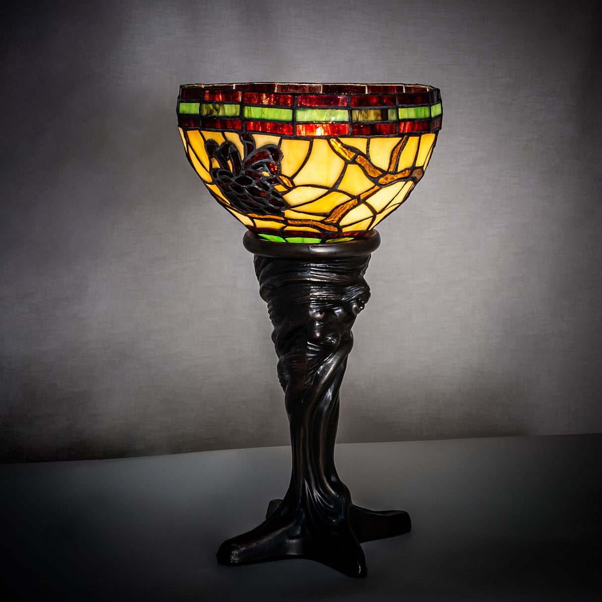 Lamp glowing in a dimly lit setting, highlighting its intricate stained-glass pinecone and branch details.
