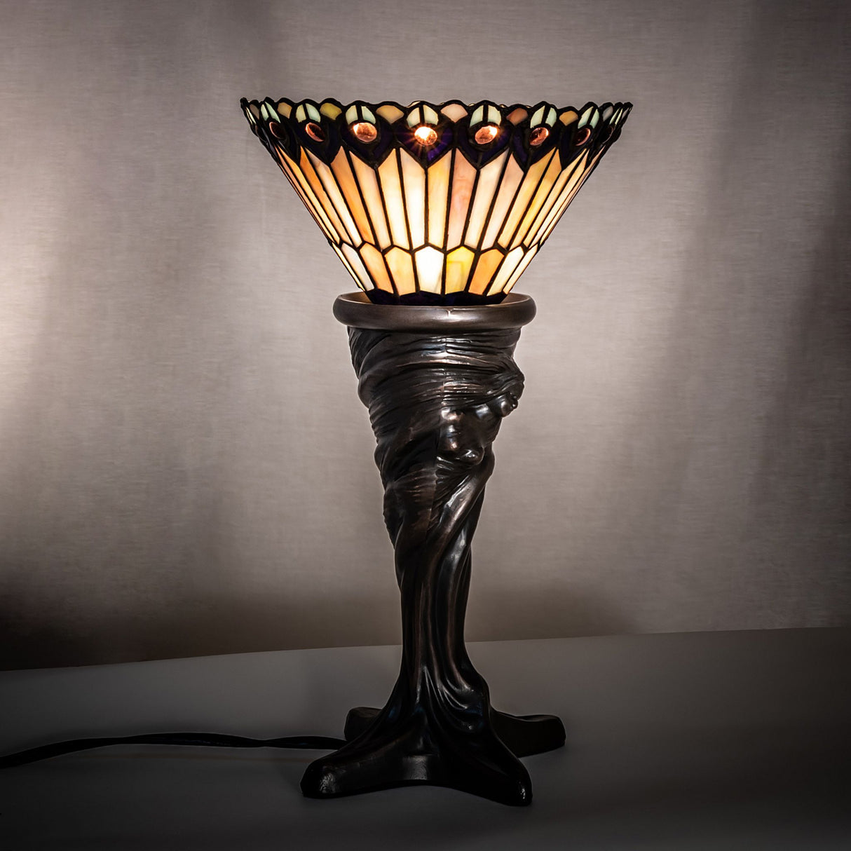 Tiffany Jeweled Peacock Mini Lamp illuminated, showcasing its colorful glass shade in a dark room.