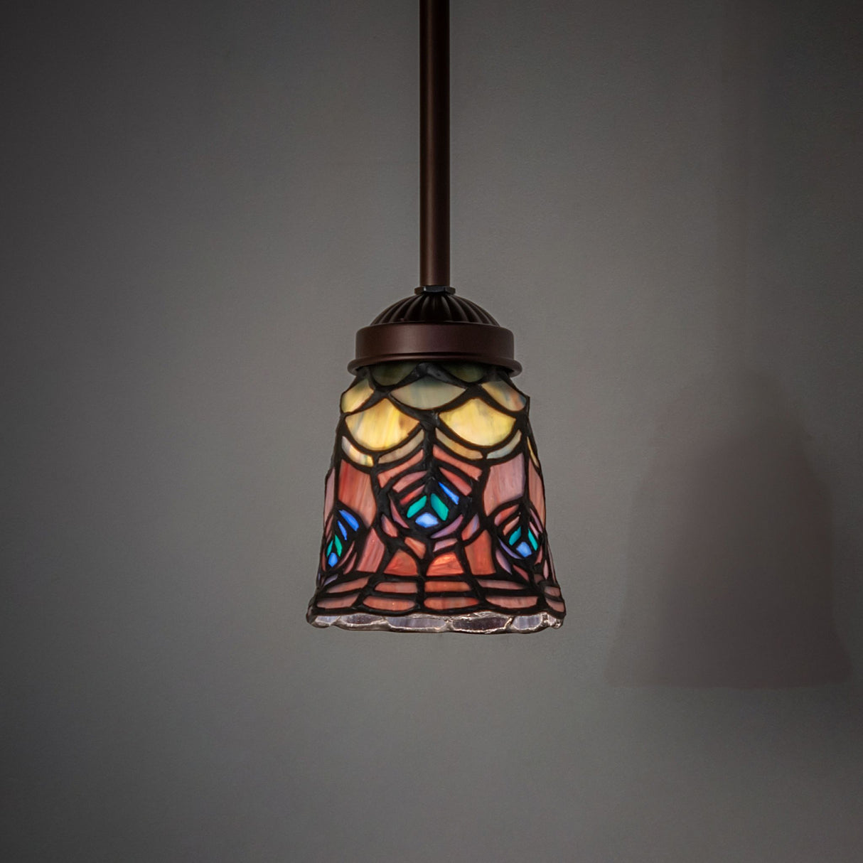 Illuminated Tiffany peacock feather mini pendant hanging in a dim setting.